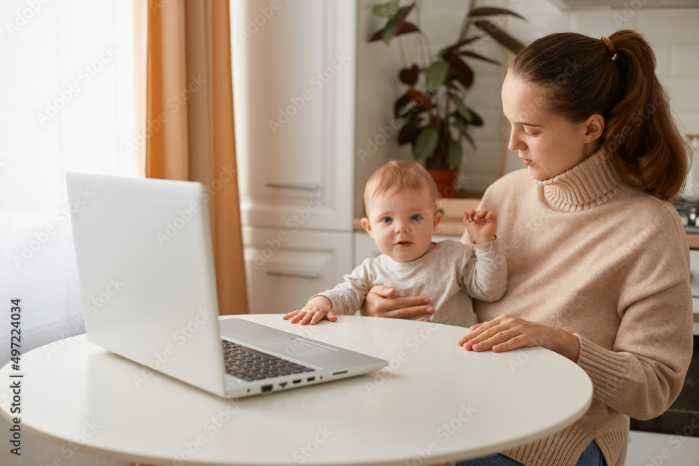 Portrait of dark haired adorable woman wearing beige jumper posing in kitchen with her infant daughter and working on laptop, looking after baby during maternity leave, working online. Portrait of dark haired adorable woman wearing beige jumper posing in kitchen with her infant daughter and working on laptop, looking after baby during maternity leave, working online.
