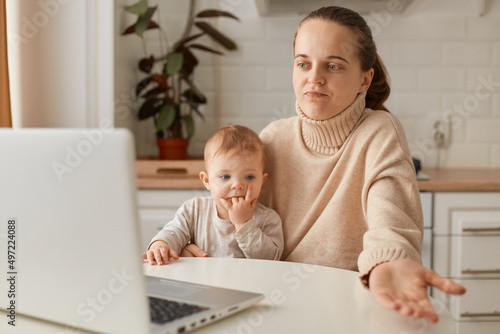 Image of puzzled dark haired woman with ponytail wearing beige jumper posing in kitchen and having online conversation, holding toddler daughter in hands, shrugging, being not sure.