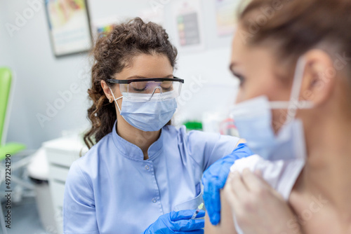 Doctor holding syringe making covid 19 vaccination injection dose in shoulder of female patient wearing mask. Flu influenza vaccine clinical trials concept, corona virus treatment, close up view.