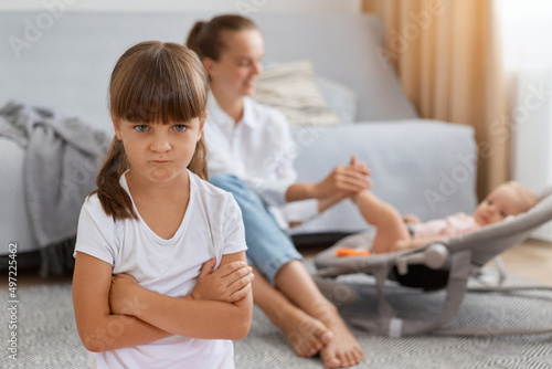 Schilderij op canvas Portrait of offended sad little girl standing with folded hands, looking at camera with sadness, jealous mother to baby sister, woman in white shirt and jeans with kid in rocking chair on background