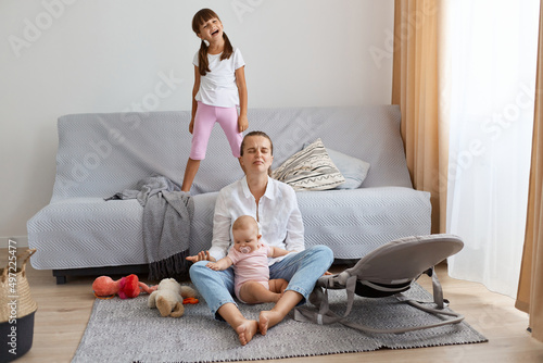 Fotografia Full length portrait of sad desperate woman wearing white shirt and jeans sitting on floor with her daughters, being depressed and tired looking after noisy kids