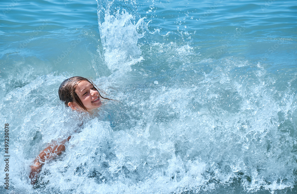 Joyful child swims in sea. Young girl is covered by wave, smiles, rejoices, bathes, swims, splashes in sea waves.
