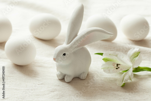 Small white bunny on light rustic background with white flowers around, close-up. Happy easter concept