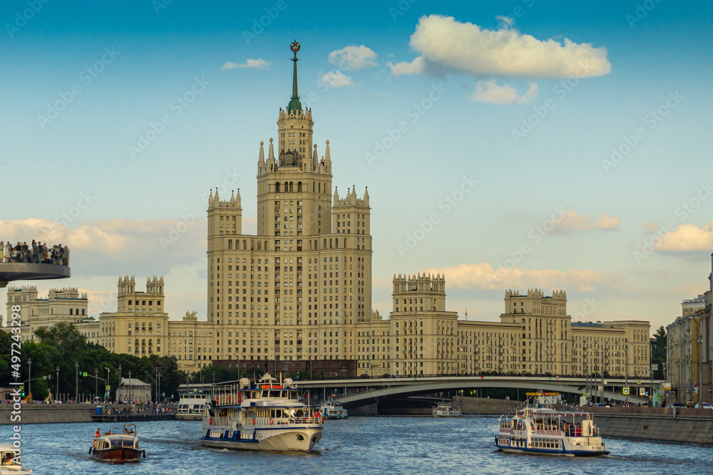 Moscow, Russia - June 16, 2019: Stalin skyscraper on background of blue sky. Pleasure boats float along Moscow River in direction of Stalinist skyscraper. People on floating bridge take pictures.