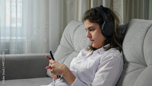 Disabled woman with infantile cerebral paralysis in headphones listens to music singing alone and holding phone sitting on sofa at home closeup.