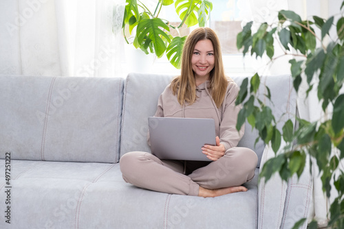 Young or middle age woman sitting with laptop on grey couch in home office with monstera plant. Concept of remote workplace and working at home.