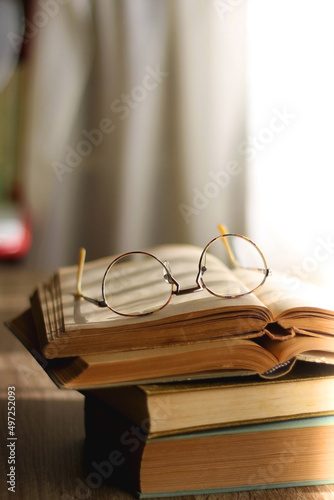 Open book and reading glasses on the table, illuminated by sunlight. Stack of vintage books in the background. Selective focus.