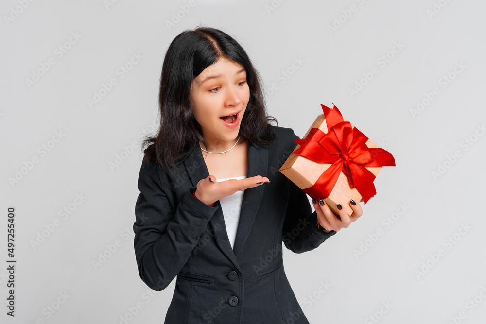 Portrait of an curious brunette girl in black blazer pointing palm of hand at a gift box, focus, intrigued what inside, standing grey background. Young emotional woman Portrait of an curious brunette girl in black blazer pointing palm of hand at a gift box, focus, intrigued what inside, standing grey background. Young emotional woman