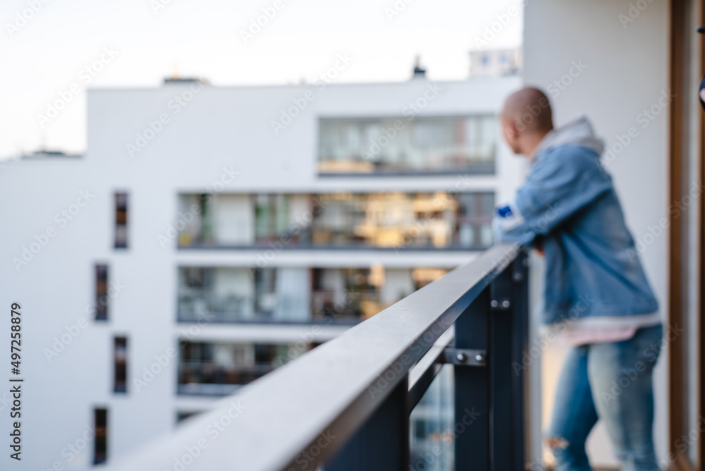 Blurred photo of a young bald person standing on the balcony, bracing their arms on a guardrail, and looking at the neighborhood. High quality photo Blurred photo of a young bald person standing on the balcony, bracing their arms on a guardrail, and looking at the neighborhood. High quality photo