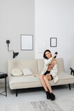 A girl sits on the couch in her home clothes and holds a therapeutic percussive vibrating massage gun in her hand. Sports recovery concept after a workout. 