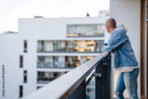 Blurred photo of a young bald person standing on the balcony, bracing their arms on a guardrail, and looking at the neighborhood. High quality photo