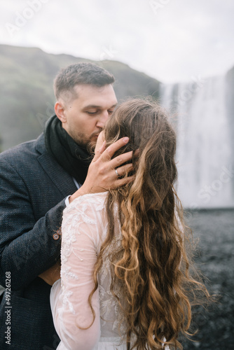 The groom is kissing his bride while droplets of Skogafoss are dissipating in the air. Iceland