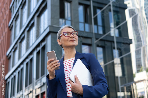 Mature asian businesswoman wearing eyeglasses using mobile phone holding laptop standing on urban street. Portrait of successful manager waiting for taxi looking away outdoors