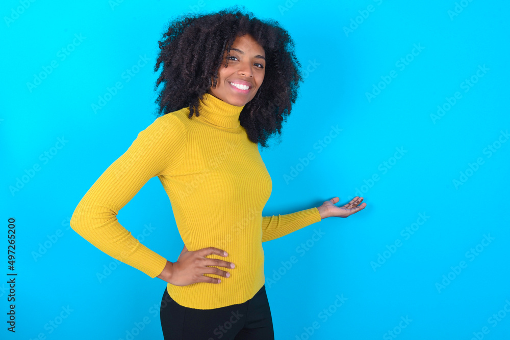 Young woman with afro hairstyle wearing yellow turtleneck over blue background feeling happy and cheerful, smiling and welcoming you, inviting you in with a friendly gesture Young woman with afro hairstyle wearing yellow turtleneck over blue background feeling happy and cheerful, smiling and welcoming you, inviting you in with a friendly gesture