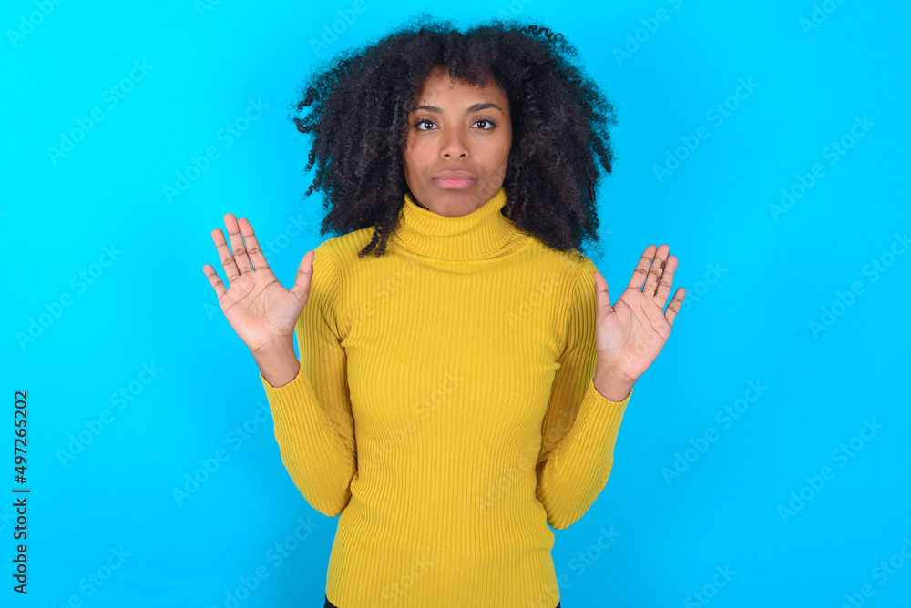 Serious Young woman with afro hairstyle wearing yellow turtleneck over blue background pulls palms towards camera, makes stop gesture, asks to control your emotions and not be nervous Serious Young woman with afro hairstyle wearing yellow turtleneck over blue background pulls palms towards camera, makes stop gesture, asks to control your emotions and not be nervous
