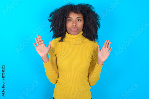 Serious Young woman with afro hairstyle wearing yellow turtleneck over blue background pulls palms towards camera, makes stop gesture, asks to control your emotions and not be nervous