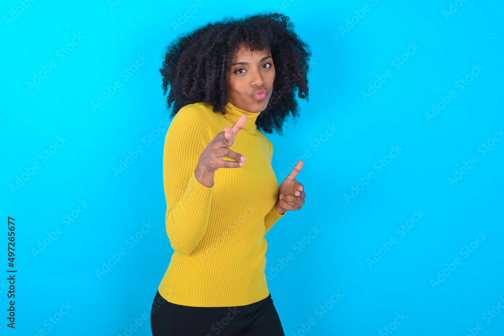 Joyful Young woman with afro hairstyle wearing yellow turtleneck over blue background wink and points index fingers at camera, chooses someone, makes finger gun pistol. Joyful Young woman with afro hairstyle wearing yellow turtleneck over blue background wink and points index fingers at camera, chooses someone, makes finger gun pistol.
