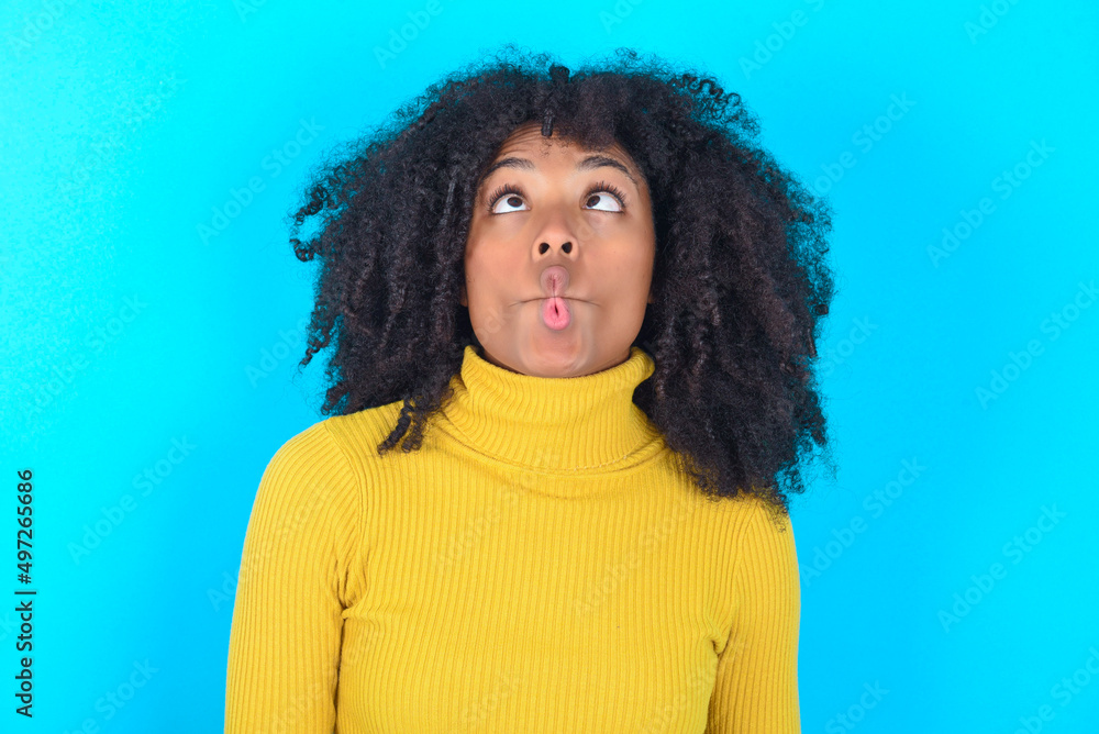 Young woman with afro hairstyle wearing yellow turtleneck over blue background making fish face with lips, crazy and comical gesture. Funny expression. Young woman with afro hairstyle wearing yellow turtleneck over blue background making fish face with lips, crazy and comical gesture. Funny expression.