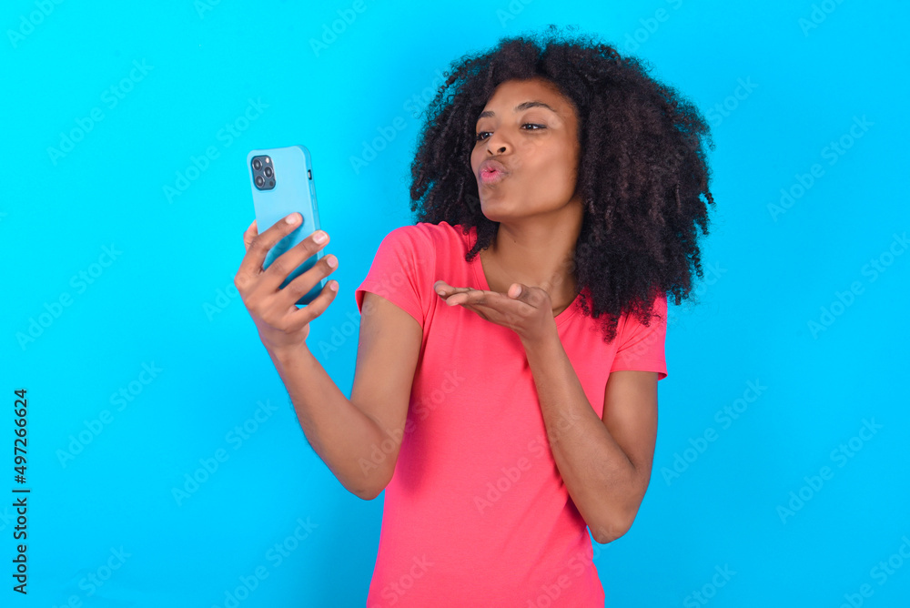 Young girl with afro hairstyle wearing pink t-shirt over blue background blows air kiss at camera of smartphone and takes selfie, sends mwah via online call. Young girl with afro hairstyle wearing pink t-shirt over blue background blows air kiss at camera of smartphone and takes selfie, sends mwah via online call.
