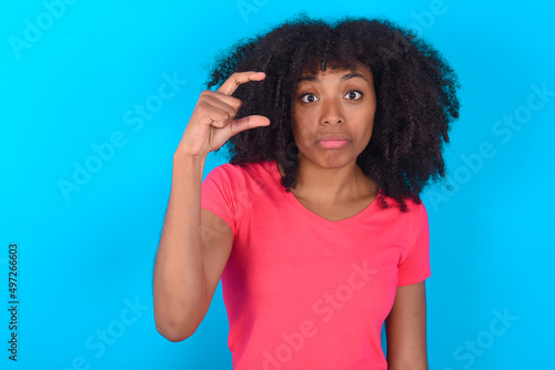Young girl with afro hairstyle wearing pink t-shirt over blue background purses lip and gestures with hand, shows something very little.