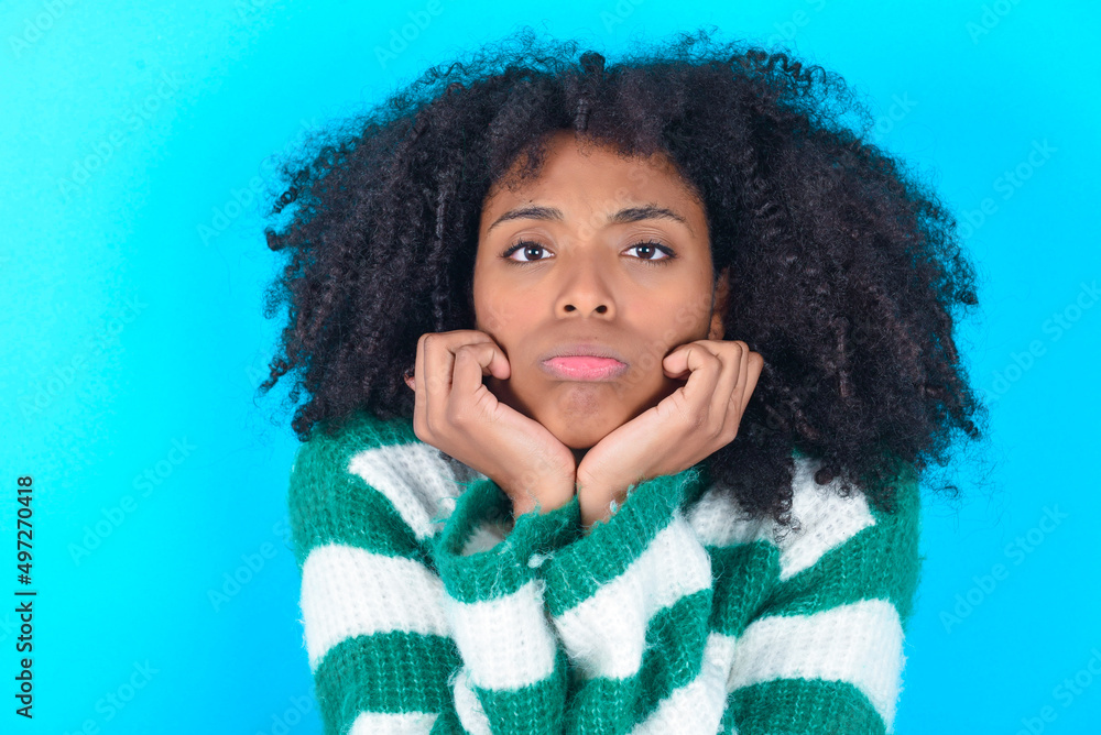 Portrait of sad Young woman with afro hairstyle wearing striped sweater over blue background hands face Portrait of sad Young woman with afro hairstyle wearing striped sweater over blue background hands face