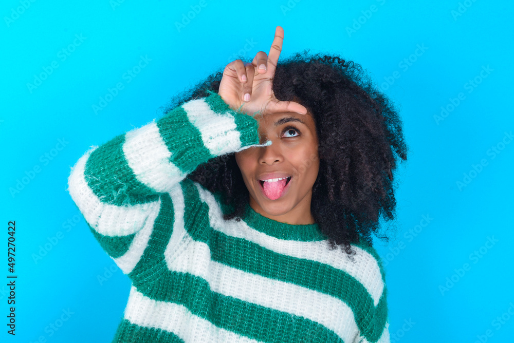 Φωτογραφία Funny Young woman with afro hairstyle wearing striped sweater over blue background makes loser gesture mocking at someone sticks out tongue making grimace face