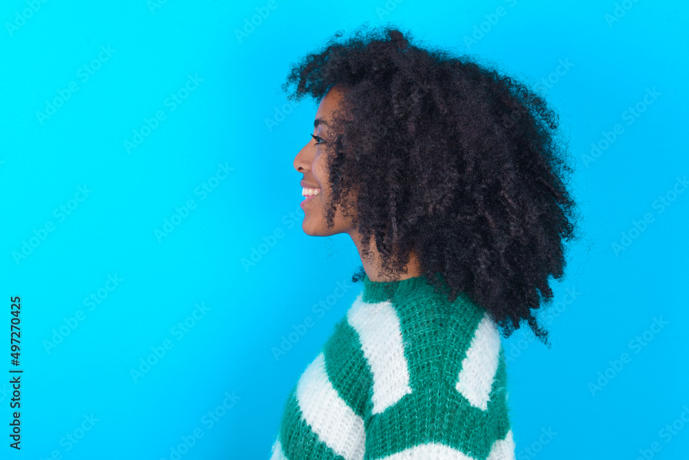 Profile portrait of nice Young woman with afro hairstyle wearing striped sweater over blue background look empty space toothy smile Profile portrait of nice Young woman with afro hairstyle wearing striped sweater over blue background look empty space toothy smile