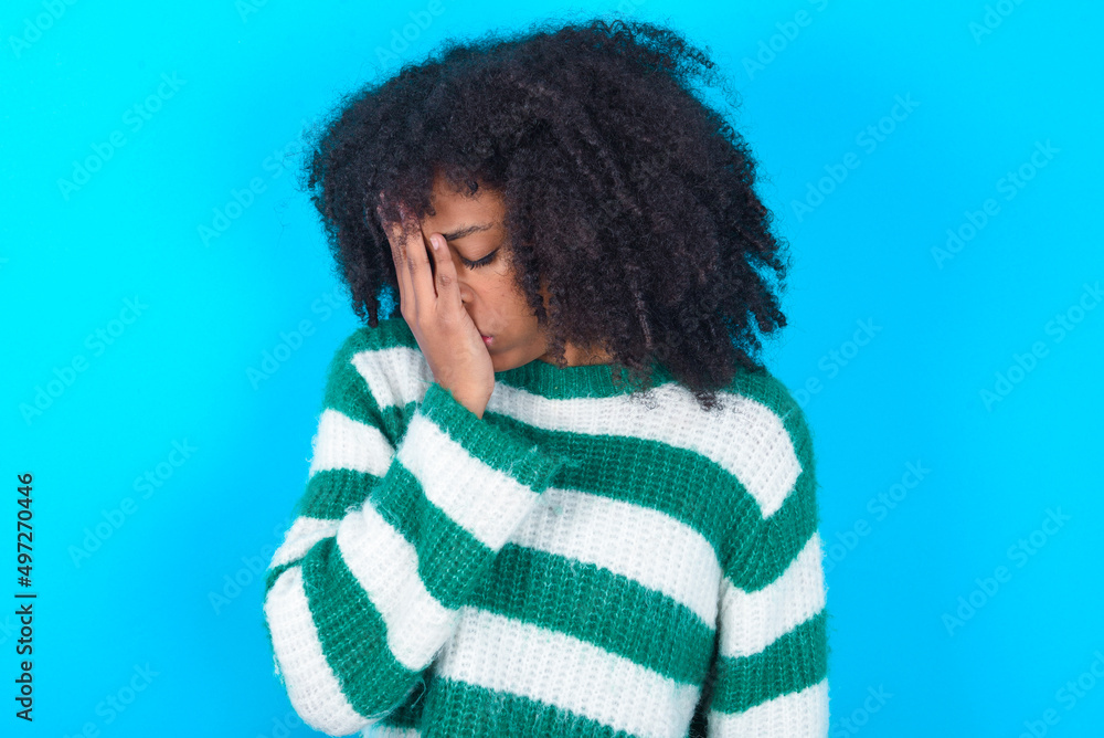 Young woman with afro hairstyle wearing striped sweater over blue background with sad expression covering face with hands while crying. Depression concept. Young woman with afro hairstyle wearing striped sweater over blue background with sad expression covering face with hands while crying. Depression concept.