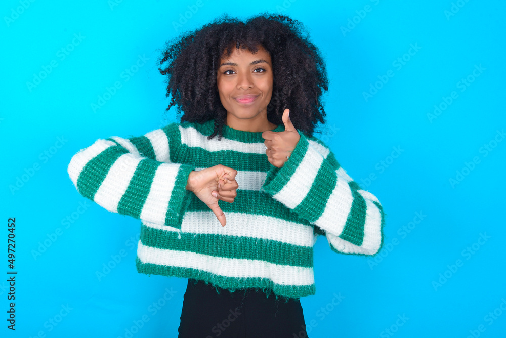 Young woman with afro hairstyle wearing striped sweater over blue background showing thumb up down sign Young woman with afro hairstyle wearing striped sweater over blue background showing thumb up down sign