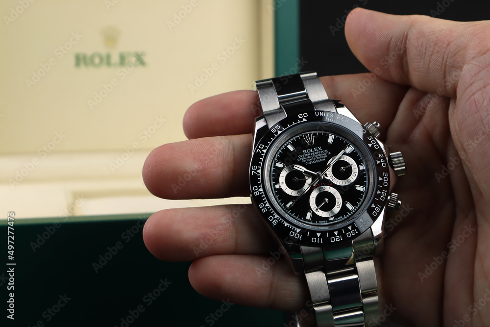 Rolex wristwatch model cosmograph daytona oyster perpetual superlative chronometer black bezel stainless steel body is in right hand with background of green rolex package box Stock Photo | Adobe Stock