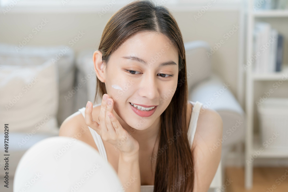 Fresh healthy skin, beautiful smile of asian young woman, girl looking at mirror, applying moisturizer on her face, putting cream treatment before makeup cosmetic routine at home. Facial Beauty. Fresh healthy skin, beautiful smile of asian young woman, girl looking at mirror, applying moisturizer on her face, putting cream treatment before makeup cosmetic routine at home. Facial Beauty.