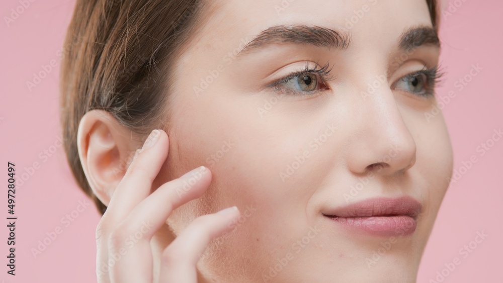 Extreme close-up of young brown-haired Caucasian woman who touches her smooth face skin, looks aside and smiles on pink background | Face care commercial concept