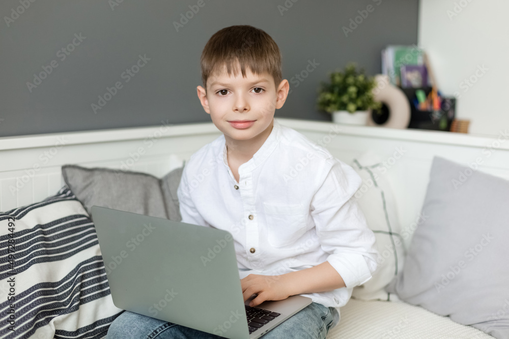 Smiling little European boy with headphones to study online using a laptop at home, cute happy little kid with headphones takes an internet lesson or a class on the computer.