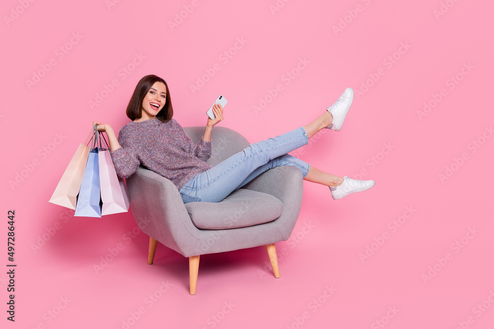Portrait of attractive cheerful girl in armchair using device holding bags order delivery isolated over pink pastel color background Portrait of attractive cheerful girl in armchair using device holding bags order delivery isolated over pink pastel color background