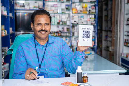 happy pharmacist showing scan here to pay qr code sign board for purchasing by looking camera at retail medical store- concept of cashless payment, wireless transaction and small business.