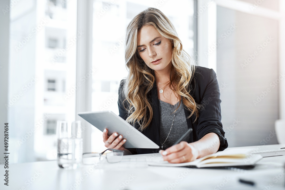 Multitasking makes it happen. Shot of a young businesswoman using a digital tablet and writing notes at her desk in a modern office. Multitasking makes it happen. Shot of a young businesswoman using a digital tablet and writing notes at her desk in a modern office.