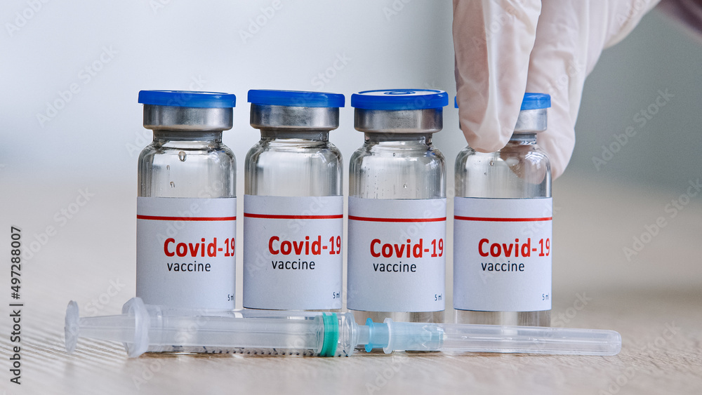 Close-up detail of four glass bottles with covid19 vaccine against coronavirus pandemic stand on wooden table in a laboratory near syringe hand in latex gloves takes one dose pharmacist sells medicine