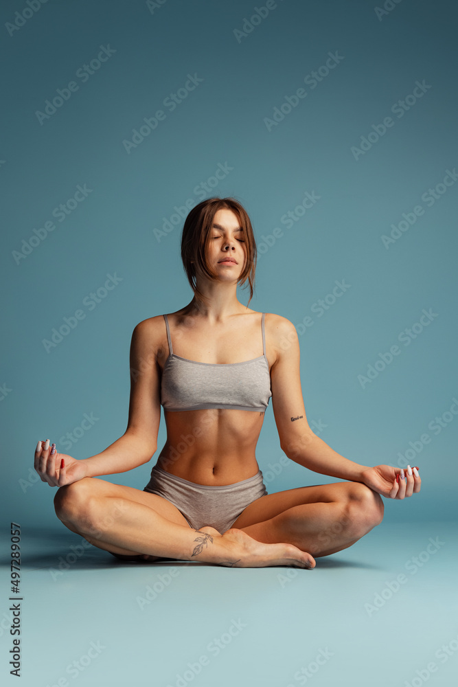 Portrait of young calm woman in underwear sitting on floor isolated over blue studio background