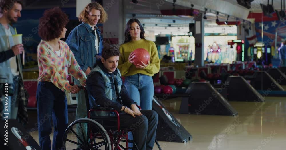 Cinematic shot of young man with disability who uses wheelchair throwing strike ball and exulting victory while having fun to play bowling together with multiracial college friends in sporting club. Cinematic shot of young man with disability who uses wheelchair throwing strike ball and exulting victory while having fun to play bowling together with multiracial college friends in sporting club.