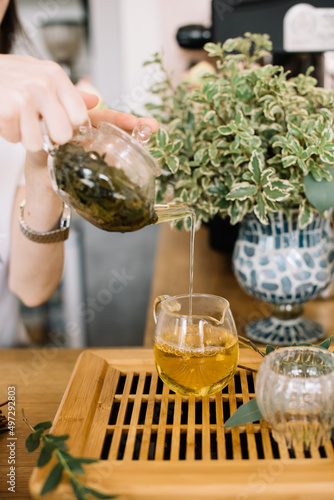 Tea ceremony is held: fresh hot oolong tea pouring through the transparent glass kettle in a matching cup, standing on the wooden tray