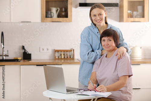 young woman teaching senior mother to use internet on laptop at home. daughter helps her elderly mother figure it out online with her personal account, teaches at modern gadget indoors