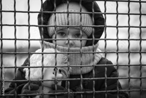 Little refugee girl with a toy behind a metal fence. Social problem of refugees and internally displaced persons. Russia's war against the Ukrainian people photo