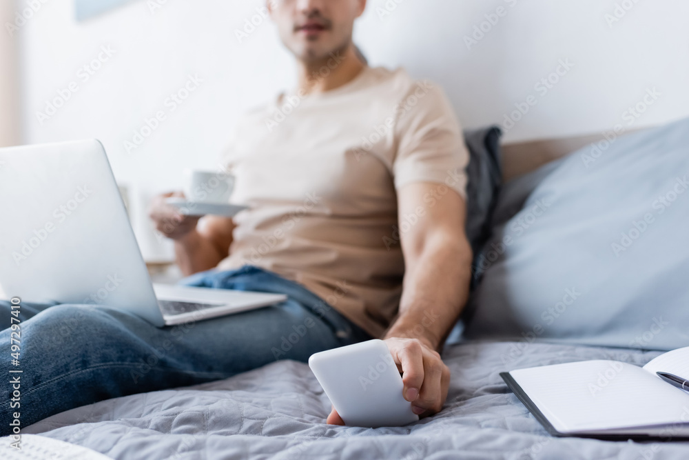 cropped view of blurred man holding cup of coffee while reaching smartphone in bedroom. cropped view of blurred man holding cup of coffee while reaching smartphone in bedroom.
