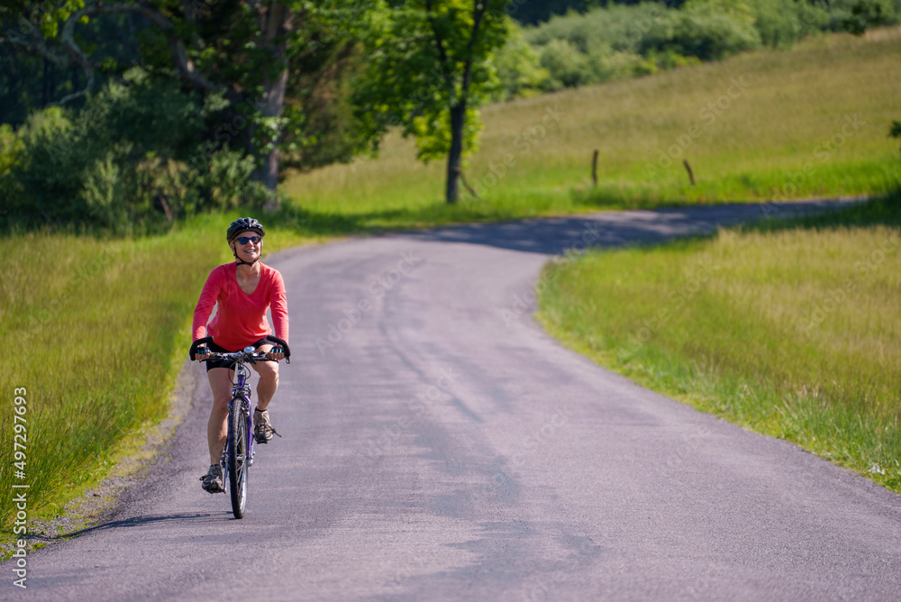 Smiling, active, mature woman wearing bike helmet and sunglasses biking on a country road on a spring summer day. Smiling, active, mature woman wearing bike helmet and sunglasses biking on a country road on a spring summer day.