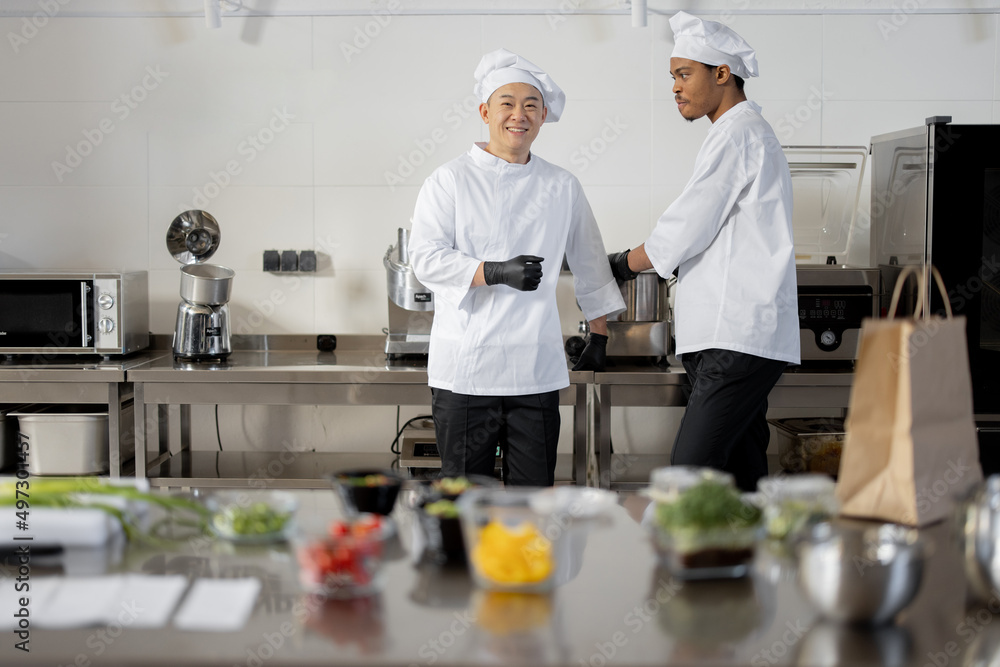 Two chef cooks standing together in professional kitchen with food ingredients in front. Latin and Asian guy working together as cooks at restaurant Two chef cooks standing together in professional kitchen with food ingredients in front. Latin and Asian guy working together as cooks at restaurant