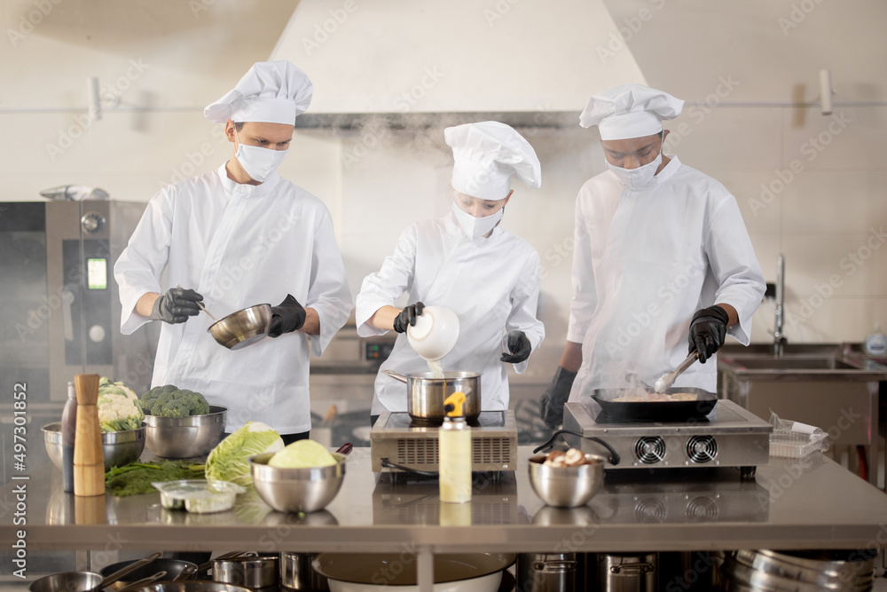 Multiracial team of cooks in uniform and face masks cooking meals for a restaurant in the kitchen. Concept of teamwork at restaurant during pandemic. Latin, Asian and European guys cooking together