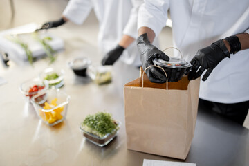 Cooks in protective gloves prepare take away food and packing them into paper bag for delivery, close-up on food. Concept of cooking food at dark kitchen for delivery