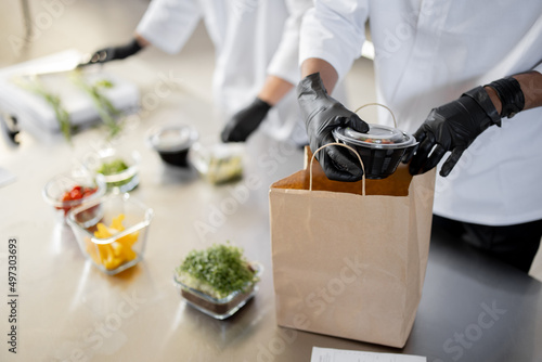 Cooks in protective gloves prepare take away food and packing them into paper bag for delivery, close-up on food. Concept of cooking food at dark kitchen for delivery photo