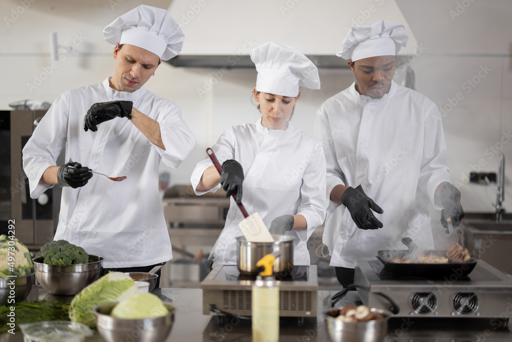 Multiracial team of three cooks in uniform cooking together in the professional kitchen. Latin cook frying meat and european guys making sauce. Concept of teamwork at restaurant