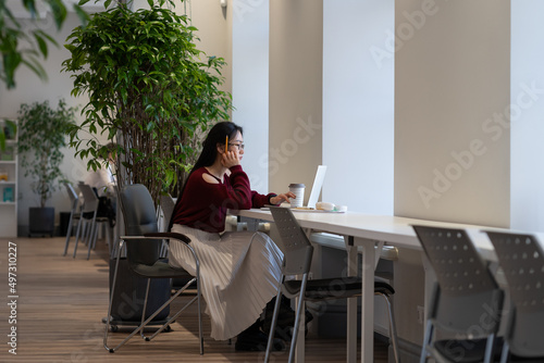 Pensive asian young woman looking thoughtfully at laptop screen while working remotely in modern public library, japanese female student using internet for educational purpose. Remote learning concept